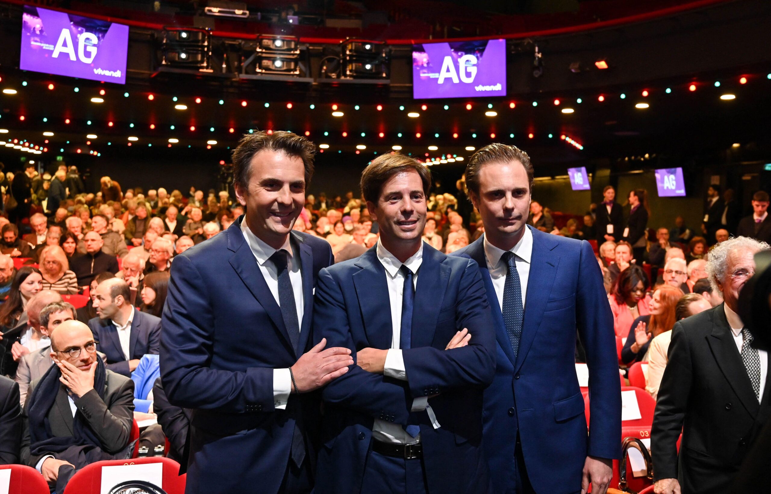 three Bollore brothers standing side by side in a large auditorium, posing for the cameras