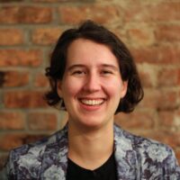 Betsy is seated in front of a brown brick wall, wearing a gray paisley blazer and plain t shirt in a professional headshot